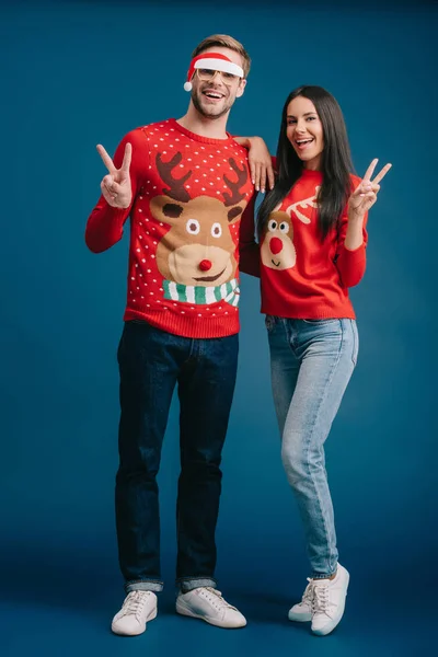 Boyfriend in glasses with santa hat and cheerful girlfriend showing victory signs isolated on blue — Stock Photo