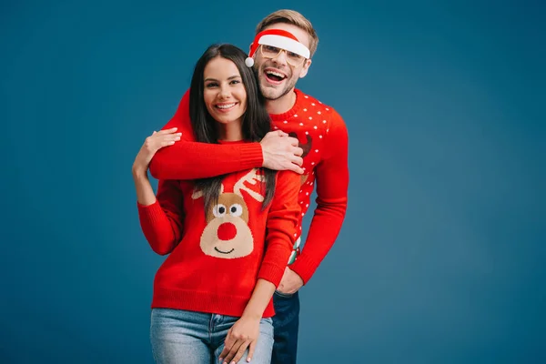 Fröhlicher Mann mit Brille und Weihnachtsmann umarmt Frau isoliert auf blauem Grund — Stockfoto