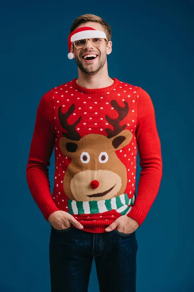 Чоловік позує в різдвяному светрі і окулярах з капелюхом Санти, ізольовані на синьому — стокове фото