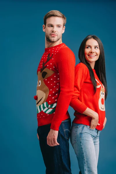 Счастливая пара в рождественских свитерах, стоящая спиной к спине, изолированная на голубом — стоковое фото
