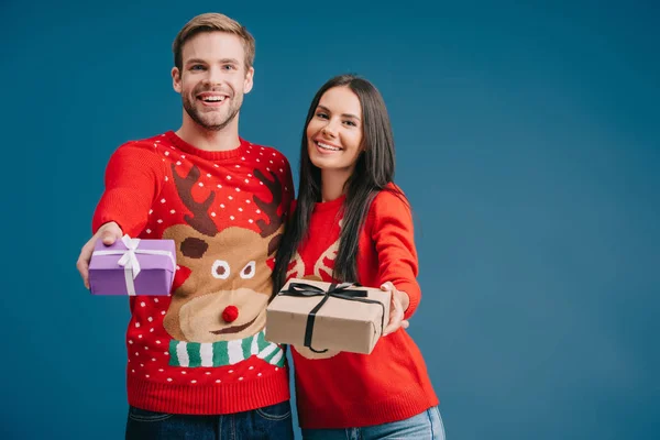 Усміхнена пара в червоних светрах тримає різдвяні подарунки ізольовані на синьому — стокове фото