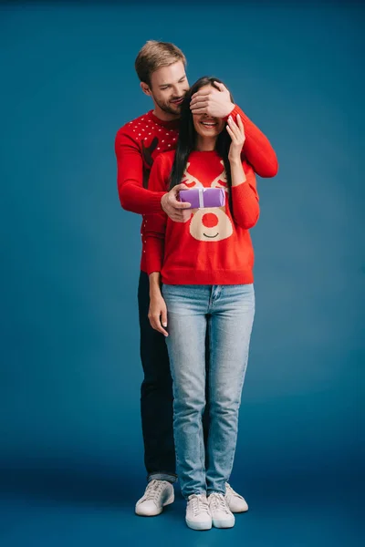 Boyfriend closing eyes of girlfriend and making christmas surprise isolated on blue — Stock Photo