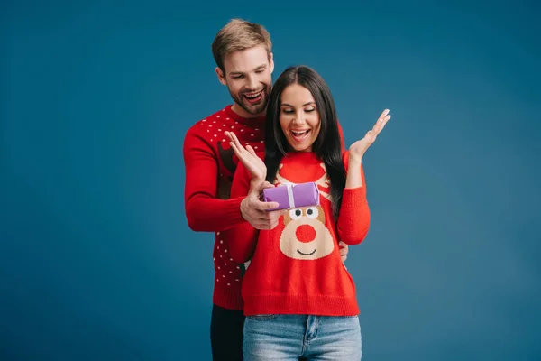 Усміхнений чоловік презентує різдвяний подарунок здивованій жінці ізольовано на синьому — стокове фото