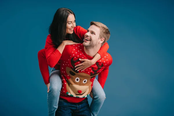 Casal alegre em camisolas de Natal piggybacking isolado em azul — Fotografia de Stock