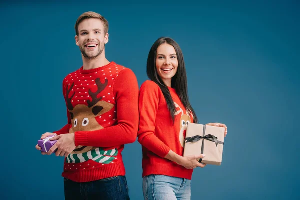 Счастливая пара в красных свитерах, держащих рождественские подарки изолированы на синий — стоковое фото