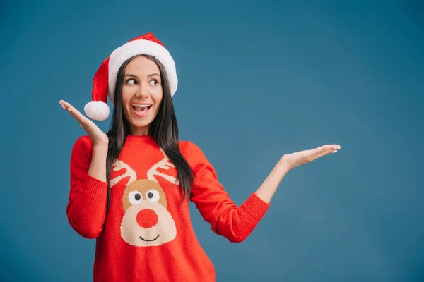 Schöne, glückliche Frau mit Weihnachtsmütze, die etwas Isoliertes auf blauem Grund zeigt — Stockfoto