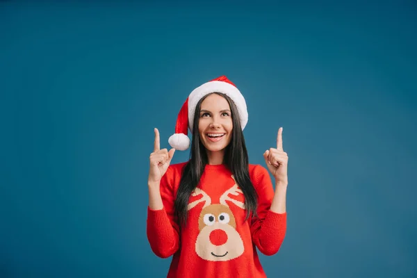 Schöne Frau mit Weihnachtsmütze, die auf etwas Isoliertes auf blauem Grund zeigt — Stockfoto