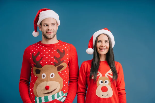 Schönes lächelndes Paar posiert in Weihnachtsmannmützen und Pullovern isoliert auf blauem Grund — Stockfoto
