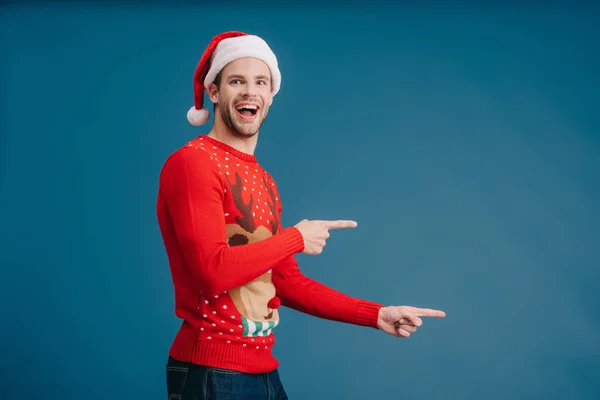 Uomo eccitato in cappello di Babbo Natale e maglione di Natale che punta isolato sul blu — Foto stock