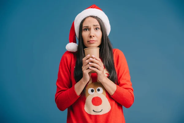 Cold girl in santa hat and christmas sweater holding coffee to go, isolated on blue — Stock Photo