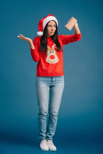 Розлючена дівчина в капелюсі Санта і різдвяний светр тримає порожню чашку кави, ізольовані на синьому — стокове фото