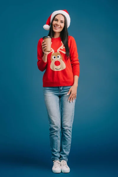 Усміхнена дівчина в капелюсі Санта і різдвяний светр тримає каву, щоб піти, ізольована на синьому — стокове фото