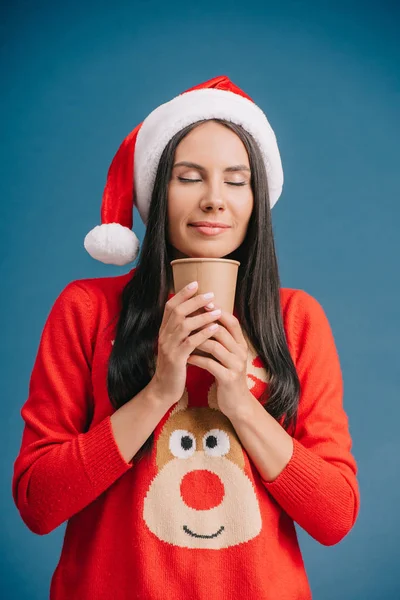 Atractiva mujer con los ojos cerrados en sombrero de santa y suéter de Navidad celebración de café para llevar, aislado en azul - foto de stock