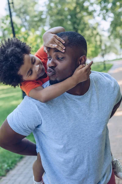 Junger afrikanisch-amerikanischer Mann huckepack und küsst entzückenden Sohn beim Spazierengehen im Park — Stockfoto