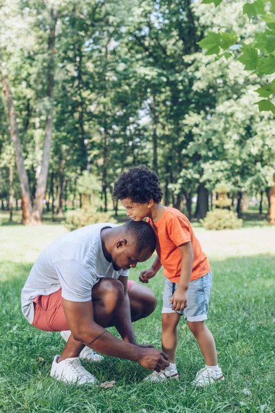 Young african american man tying shoelaces of cute son in park — Stock Photo