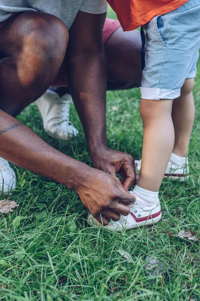 Vista parcial del hombre afroamericano atando cordones de zapatos de hijo en parque - foto de stock