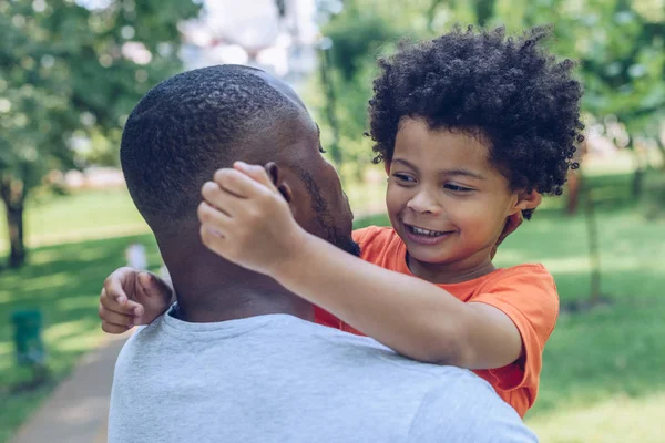 Bonito afro-americano menino abraçando pai enquanto caminhando no parque — Fotografia de Stock