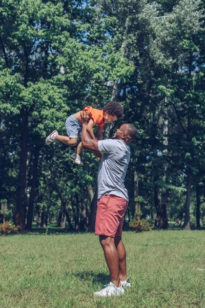 Feliz afroamericano hombre sosteniendo alegre hijo por encima de la cabeza mientras se divierten en parque - foto de stock