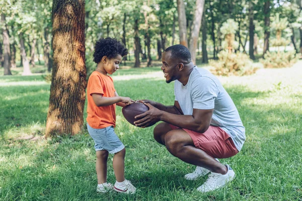 Красивый африканский американец показывает регбийный мяч очаровательному сыну в парке — стоковое фото