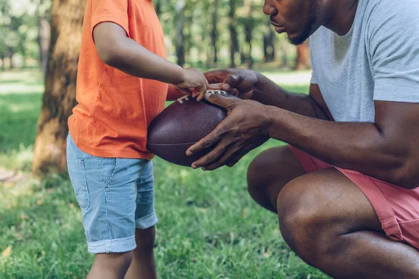 Обрезанный вид африканского американского отца, показывающего регби мяч сыну в парке — стоковое фото