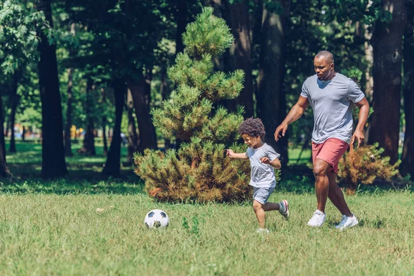 Alegre afroamericano padre e hijo jugando fútbol en parque - foto de stock