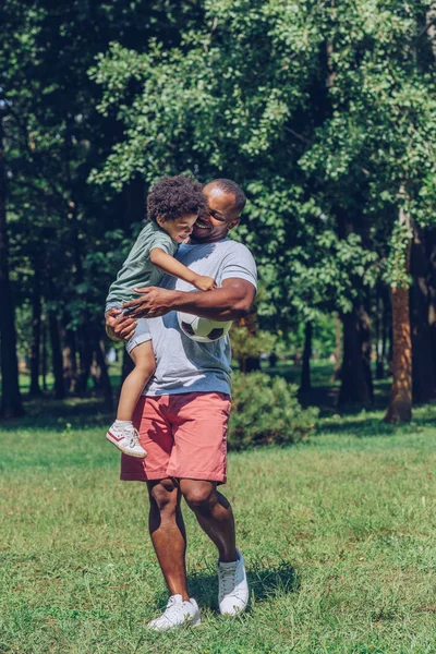 Glücklicher afrikanisch-amerikanischer Junge hält Fußballball, während er auf den Händen seines Vaters sitzt — Stockfoto
