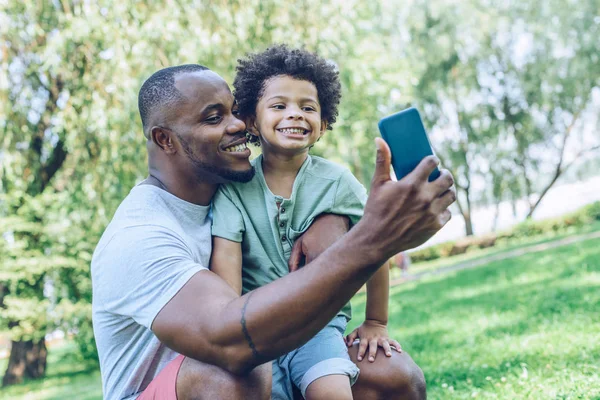 Feliz afro-americano pai e filho tomando selfie no smartphone no parque — Fotografia de Stock