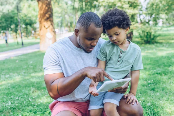 Молодой африканский отец-американец, использующий цифровой планшет, чтобы очаровательный сын в парке — стоковое фото
