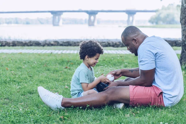 Giovane africano americano mostrando figlio guanto da baseball mentre seduto sul prato nel parco — Foto stock