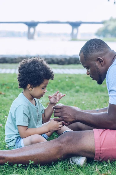 Молодой африканский американец показывает сыну бейсбольный мяч, сидя на газоне в парке — стоковое фото