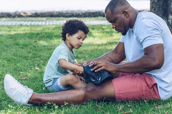 Bel homme afro-américain montrant gant de baseball à son fils assis sur la pelouse dans le parc — Photo de stock