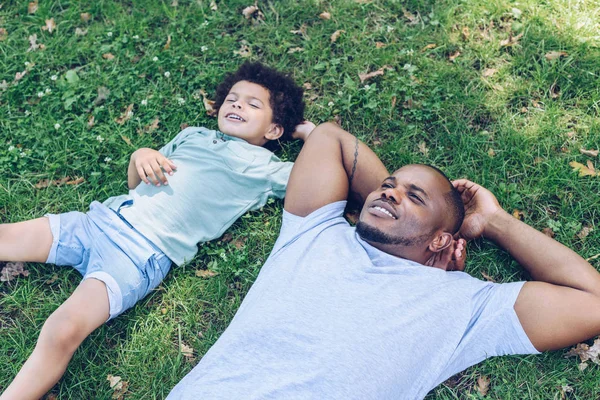 Sonriente afroamericano padre e hijo tumbado en el césped mientras descansa en el parque - foto de stock