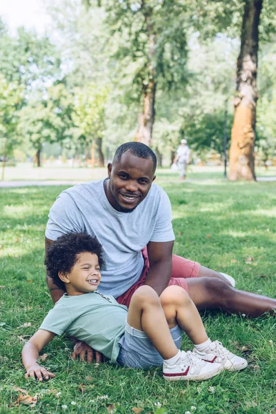 Heureux homme afro-américain souriant à la caméra tout en étant assis sur la pelouse près de son fils dans le parc — Photo de stock