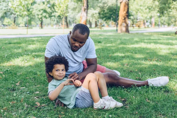 Feliz afroamericano padre e hijo sentado en el césped en el parque - foto de stock