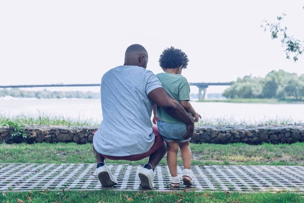 Back view of african american father and son squatting and looking at river in park — Stock Photo