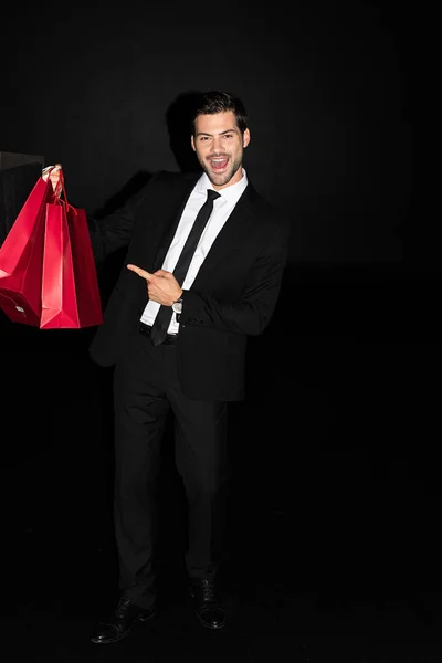 Homem animado elegante em terno mostrando sacos de compras vermelhos isolados em preto — Fotografia de Stock