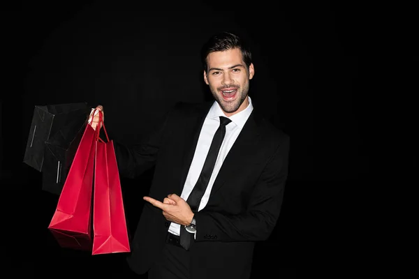 Cheerful elegant man in suit pointing at shopping bags isolated on black — Stock Photo