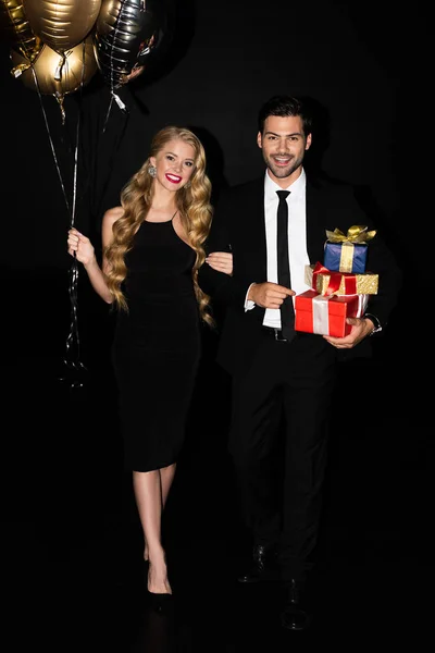 Pareja alegre con globos dorados y regalos aislados en negro - foto de stock