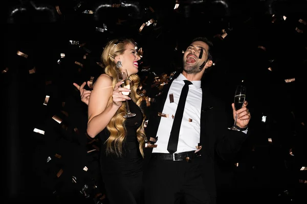 Beautiful cheerful couple celebrating with champagne on black with golden confetti — Stock Photo