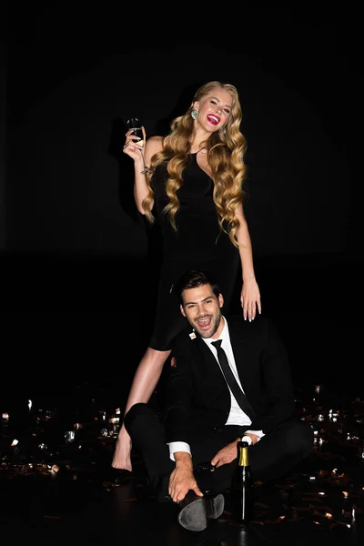 Young couple celebrating with champagne on black with golden confetti — Stock Photo