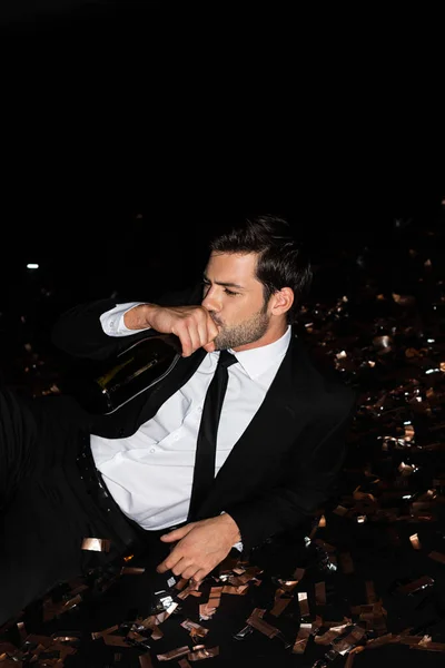 Stilvoller Mann trinkt Champagner aus Flasche, während er mit goldenem Konfetti auf dem Boden liegt — Stockfoto