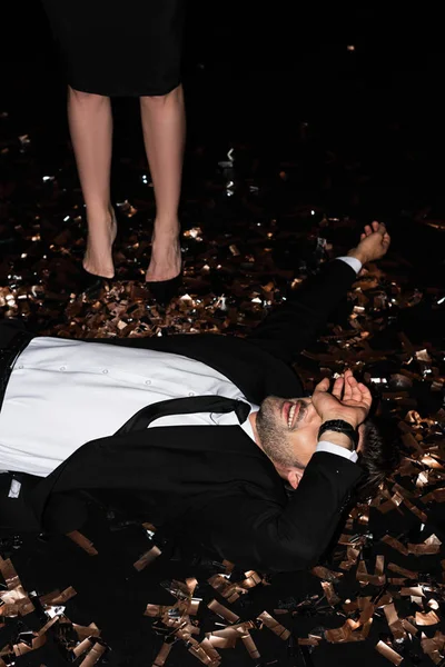 Giovane uomo sdraiato su confetti d'oro mentre la ragazza in piedi vicino su nero — Foto stock