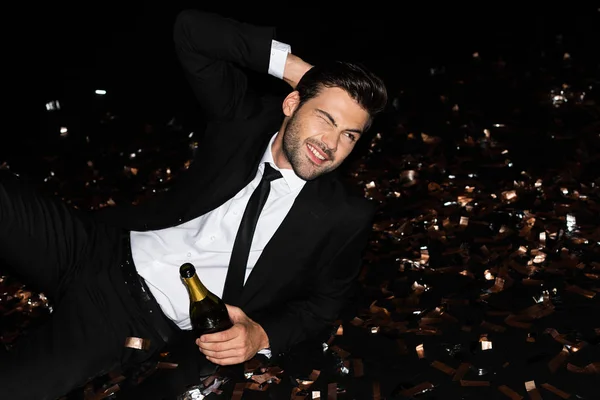 Eleganter Mann mit Champagnerflasche auf dem Boden liegend mit goldenem Konfetti für den Urlaub — Stockfoto