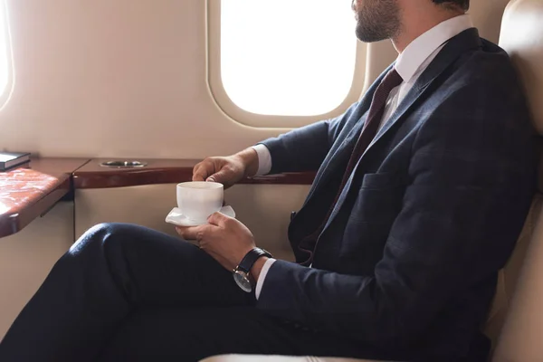 Ausgeschnittene Ansicht eines Geschäftsmannes im Anzug, der während einer Geschäftsreise eine Tasse Kaffee im Flugzeug hält — Stockfoto