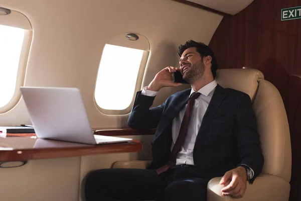 Hombre de negocios riendo hablando en el teléfono inteligente en avión con el ordenador portátil durante el viaje de negocios - foto de stock