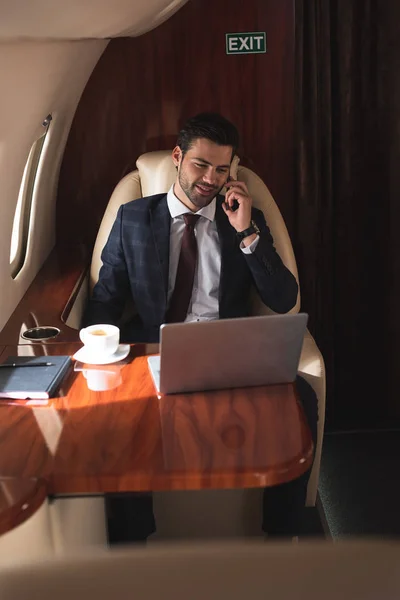 Smiling businessman talking on smartphone in plane with laptop during business trip — Stock Photo