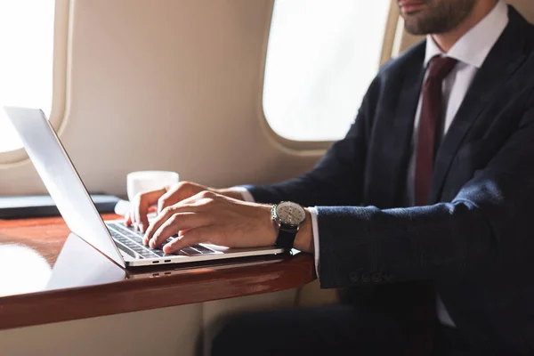 Обрезанный вид бизнесмена, печатающего на ноутбуке в самолете во время деловой поездки — стоковое фото