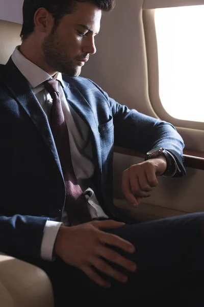 Guapo hombre de negocios mirando el reloj mientras está sentado en el avión — Stock Photo