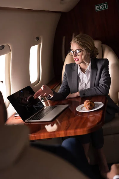 Mujer de negocios rubia trabajando en portátil en avión con croissant durante su viaje de negocios - foto de stock