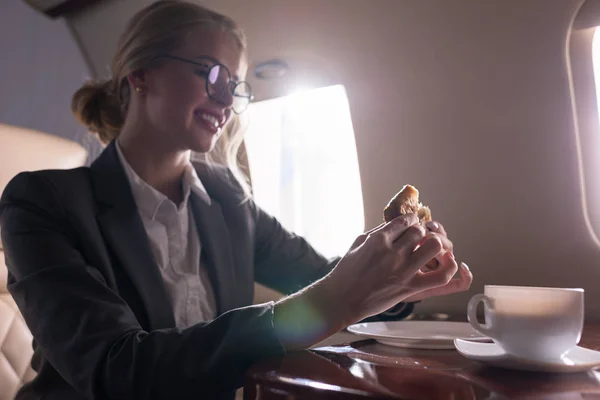 Attractive businesswoman drinking coffee with croissant in plane during business trip — Stock Photo